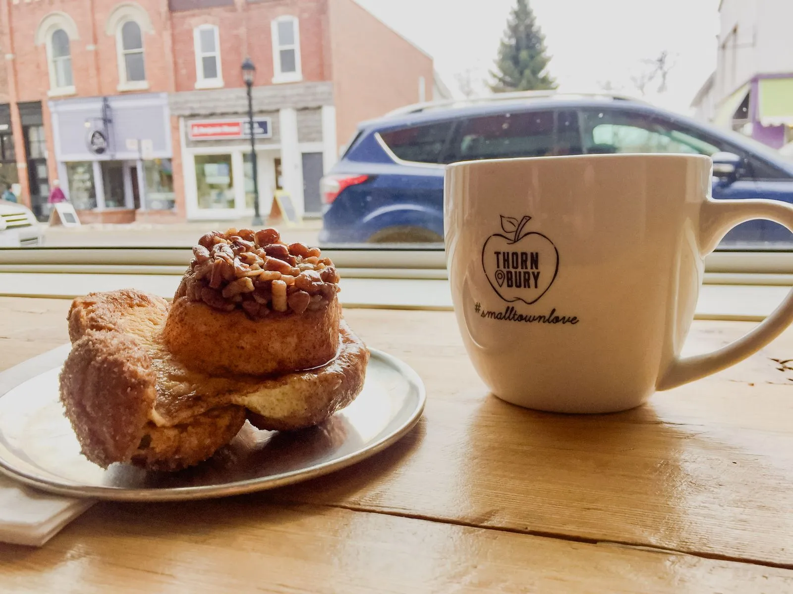 A coffee mug and sticky bun on a wooden cafe counter, with a small-town main street through the window behind